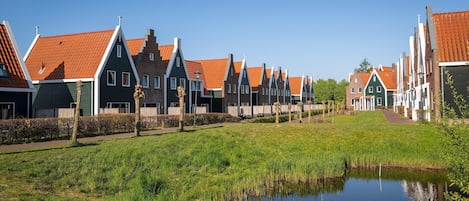 A row of traditional houses with red-tiled roofs and white trim, a grassy area in the foreground, and a small pond.