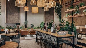 Un interior de restaurante moderno con varias mesas de comedor, lámparas colgantes de mimbre y una variedad de plantas en macetas.