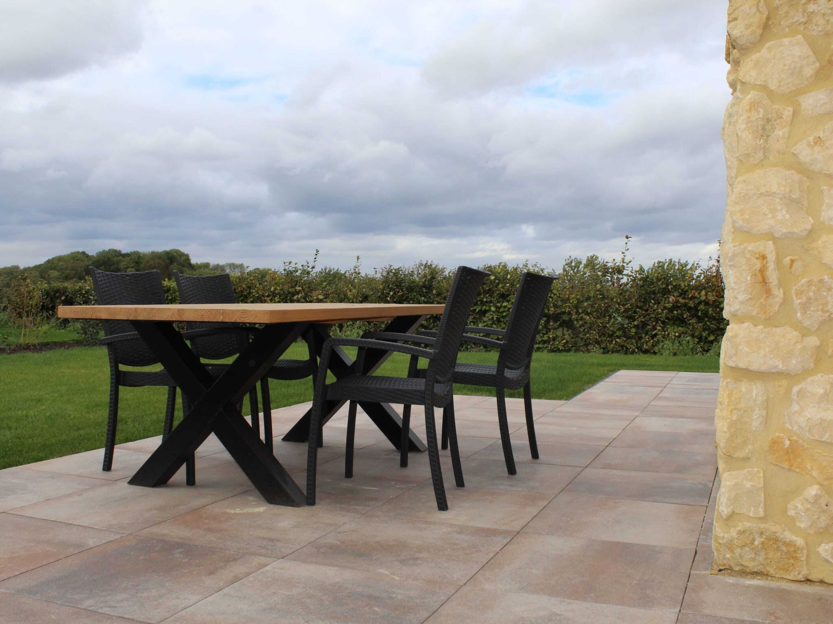 An outdoor patio with a wooden table and black chairs, surrounded by stone walls and a grassy area.