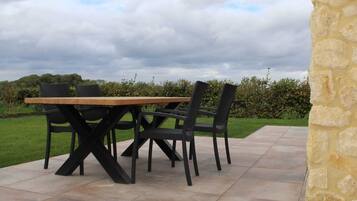 An outdoor patio with a wooden table and black chairs, surrounded by stone walls and a grassy area.