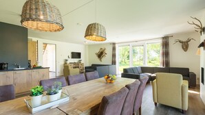 A modern living room with a wooden dining table, comfortable seating, and large windows offering a view of the outdoors.