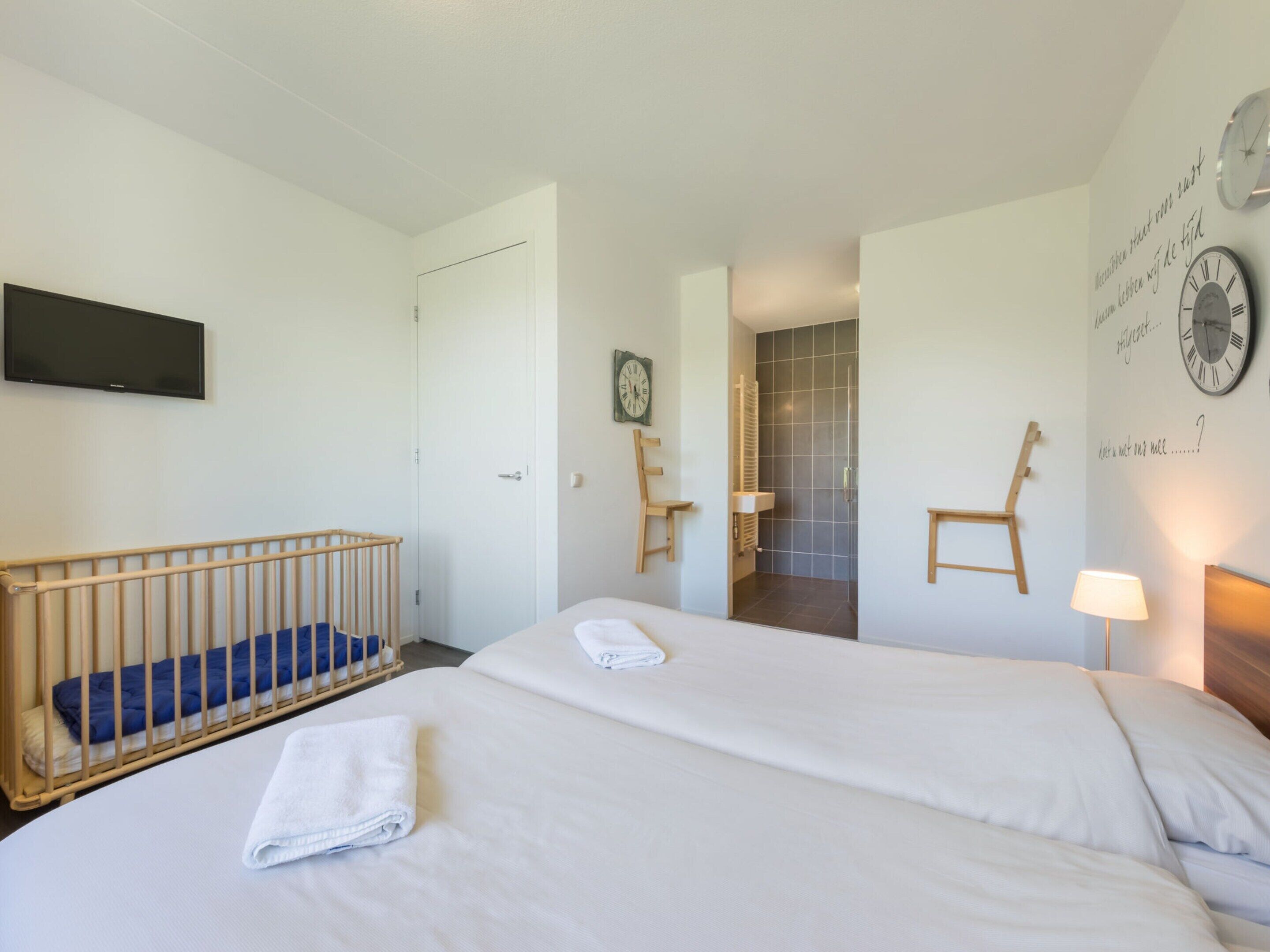 Une chambre d’hôtel avec deux lits, un berceau, une télévision, une douche et une horloge murale.
