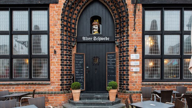 Un bâtiment en briques avec une entrée en arc noir et une enseigne indiquant « Alter Schwedes ». Deux plantes en pot sont placées sur les marches, et il y a des sièges extérieurs avec des chaises et des tables en osier.