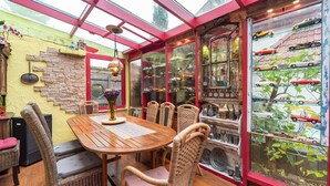 Une pièce chaleureuse avec une table en bois et des chaises en osier, une vitrine contenant des voitures miniatures et une fenêtre à cadre rouge.