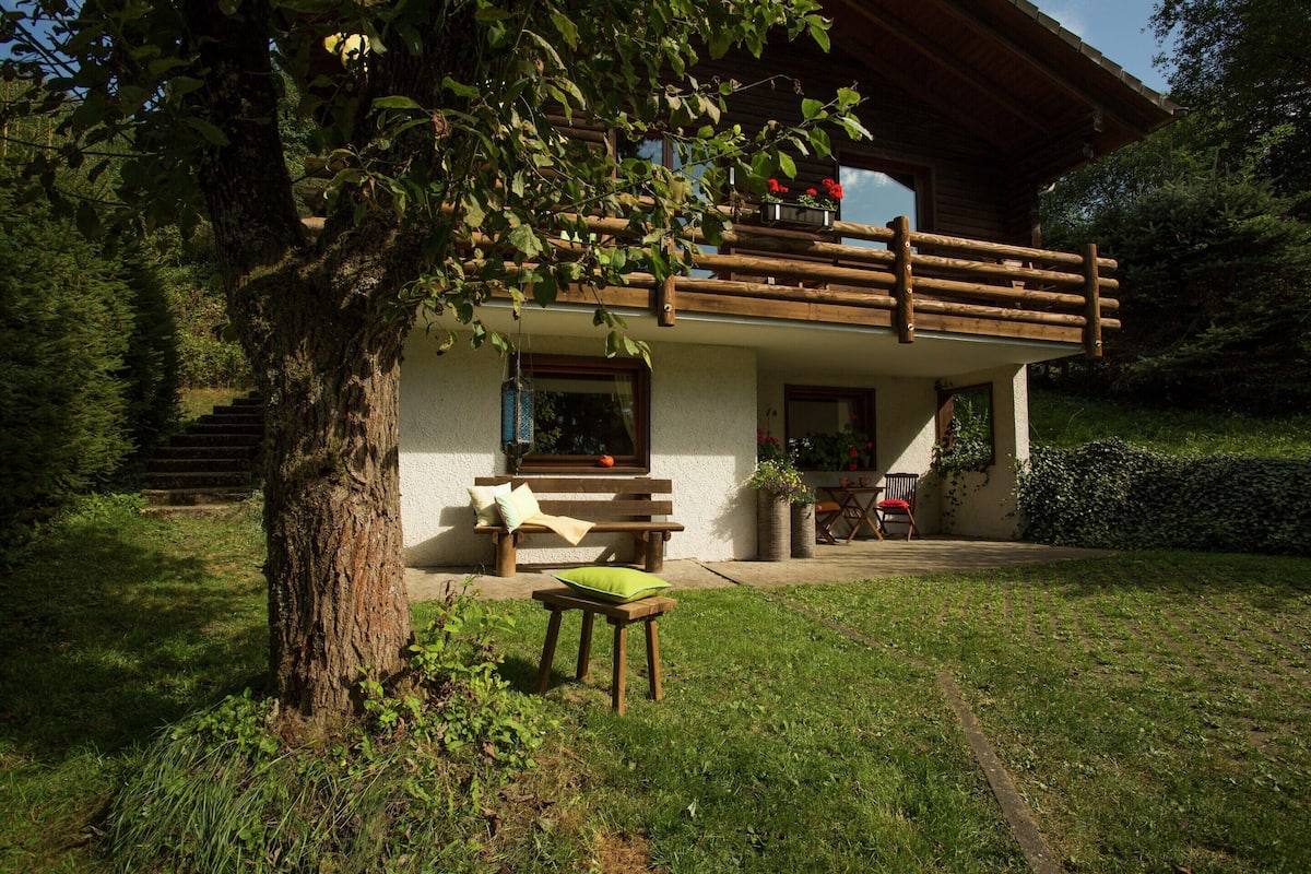 A rustic chalet with a wooden balcony, a bench, and a small table with a cushion.
