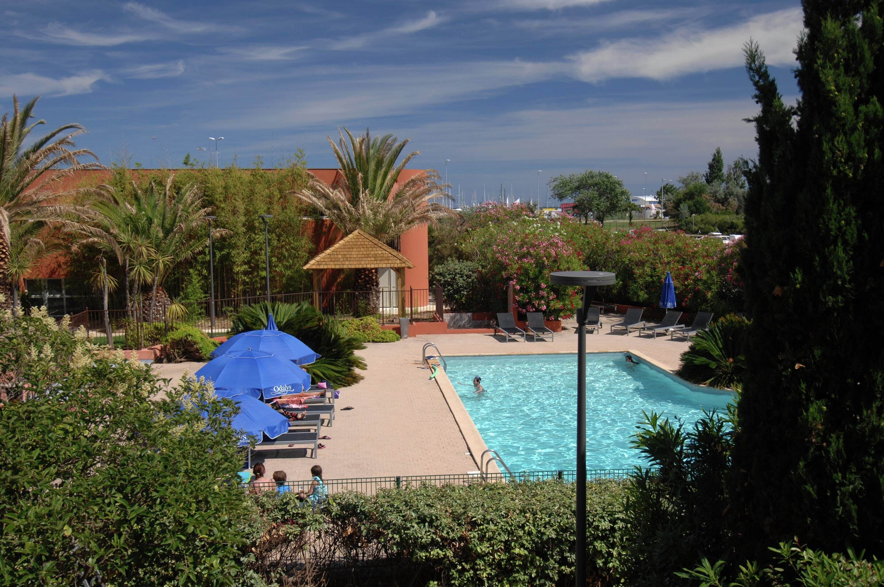 A swimming pool with lounge chairs and umbrellas, surrounded by palm trees and a building with a red roof.