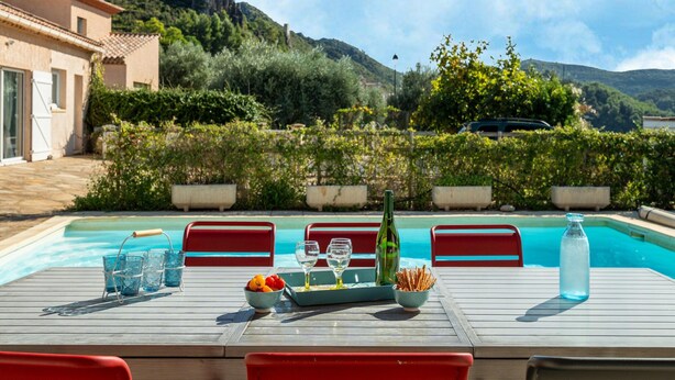Ein Tisch am Pool mit Gläsern, einer Flasche und Snacks, umgeben von roten Stühlen, mit Bergen und Pool im Hintergrund.