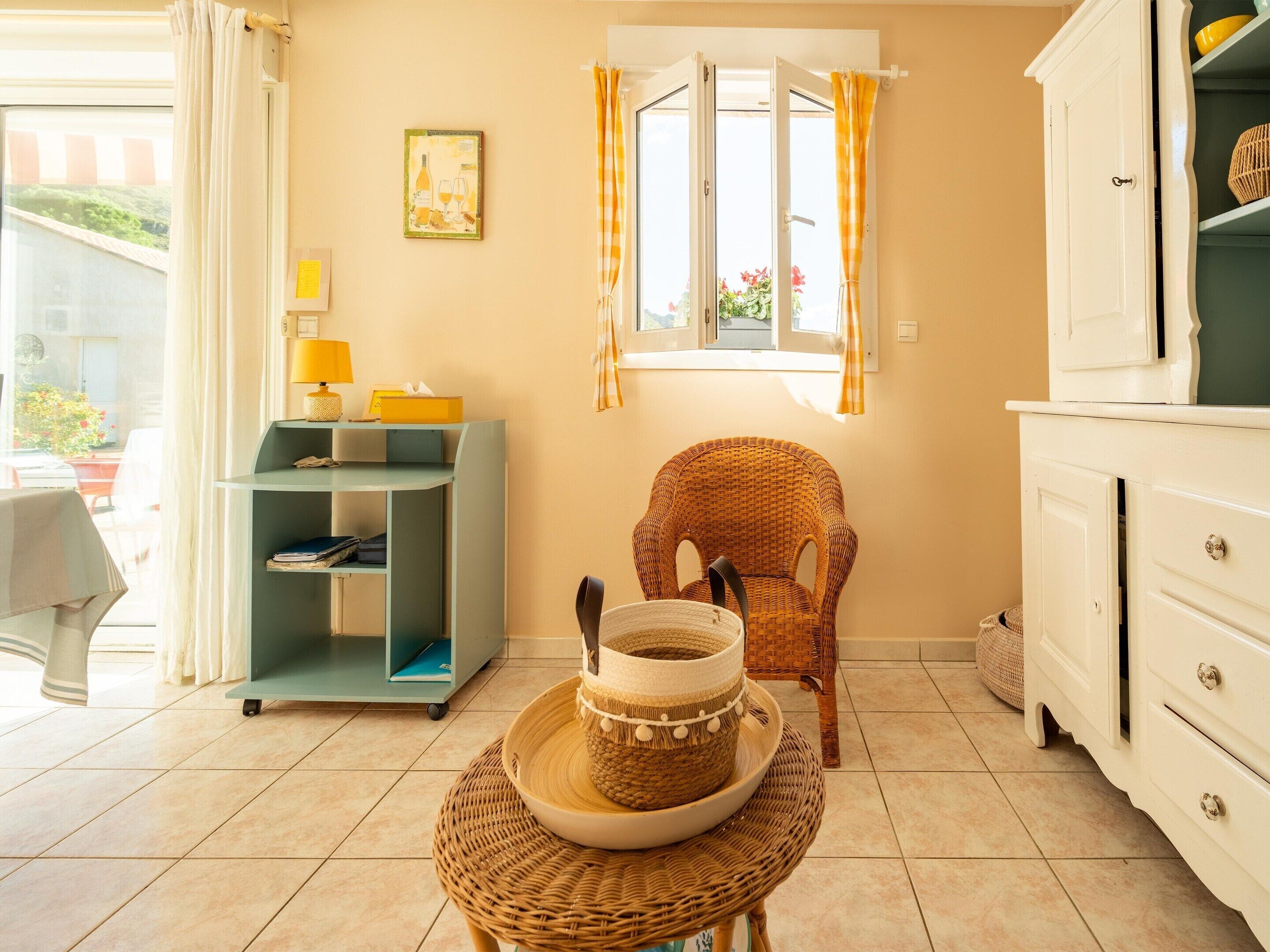 Une pièce comprenant un fauteuil en osier, une petite table avec un bol, un bureau avec une lampe et une fenêtre avec des rideaux.