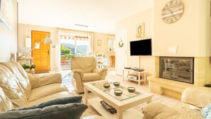 Ein Wohnzimmer mit einer Couch, einem Sessel, einem Couchtisch, einem Fernseher, einem Kamin und Blick durch eine Glastür.