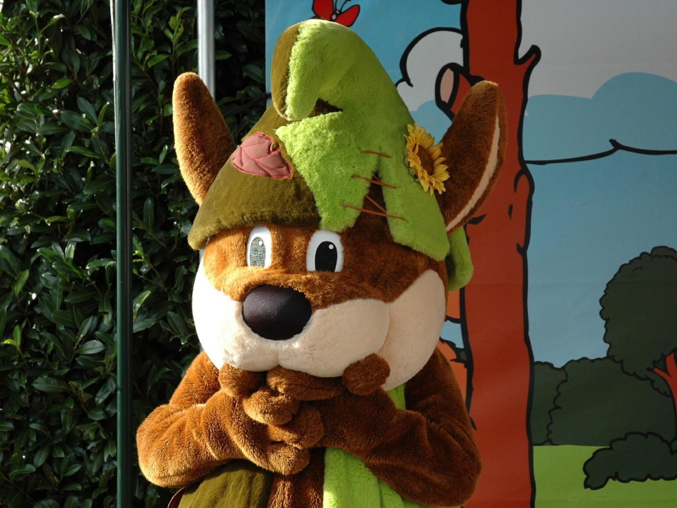 A plush toy character with a hat and a flower, standing in front of a colorful mural.