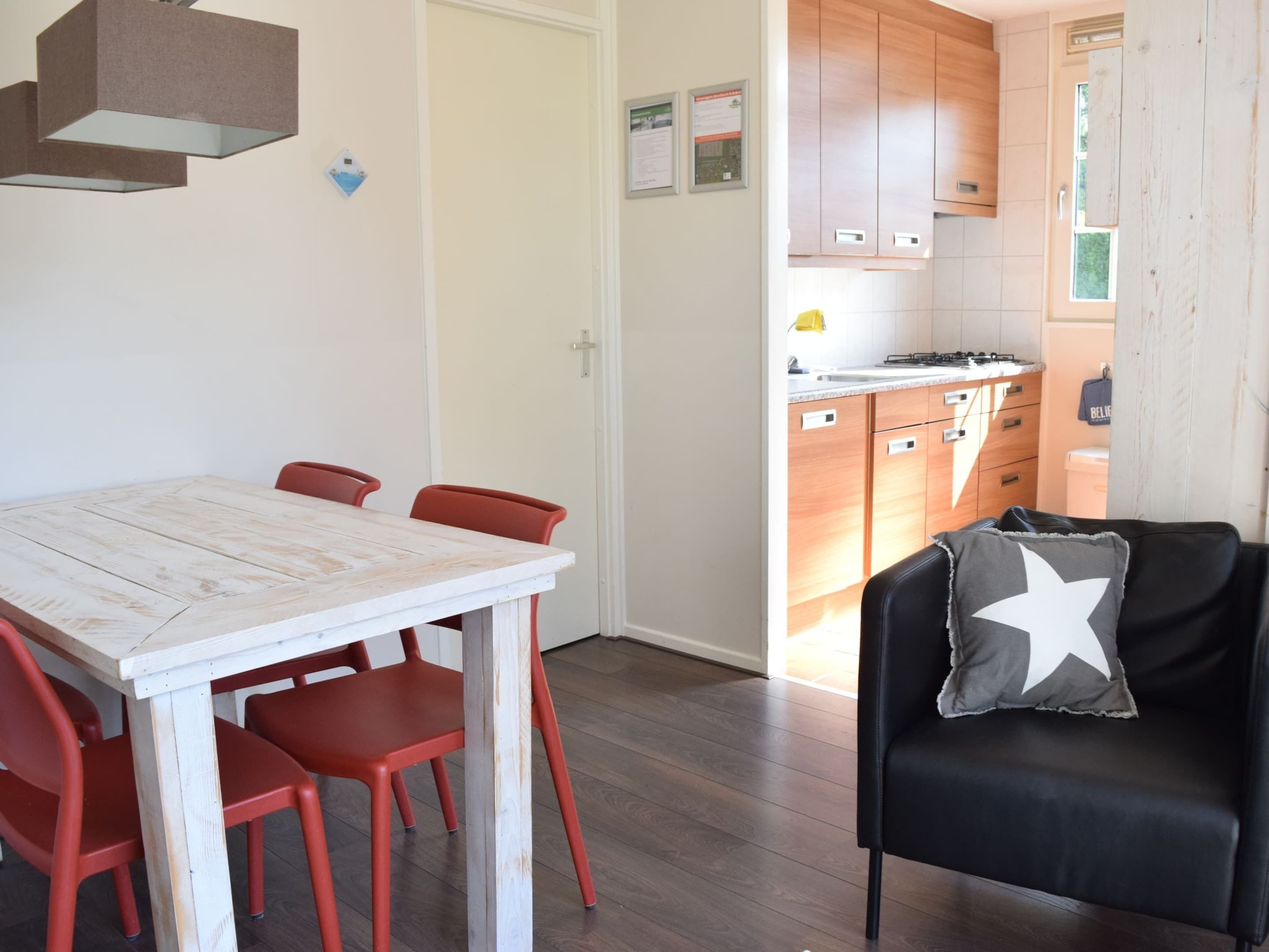Un coin repas avec une table en bois blanc et des chaises rouges, un canapé en cuir noir agrémenté d’un coussin en forme d’étoile, et des meubles en bois dans la cuisine adjacente.