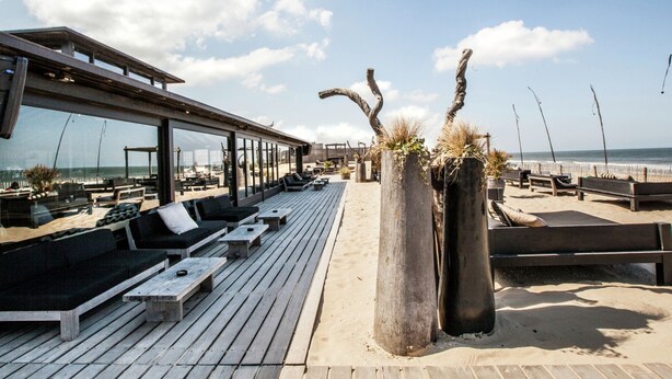 A beachside lounge with wooden benches and lounge chairs, large windows overlooking the ocean, and rustic planters with greenery. - Spacious Lodge With 2 Bathrooms, Near the Sea (Zandvoort)