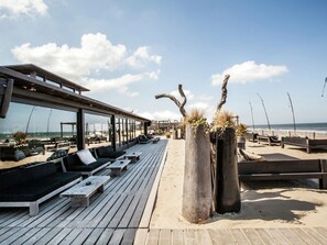 A beachside lounge with wooden benches and lounge chairs, large windows overlooking the ocean, and rustic planters with greenery. - Spacious Lodge With 2 Bathrooms, Near the Sea (Zandvoort)