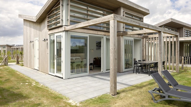 A modern house with a large glass door, a patio with a table and chairs, and a wooden deck. - Luxury Chalet Near Beach (Kamperland)