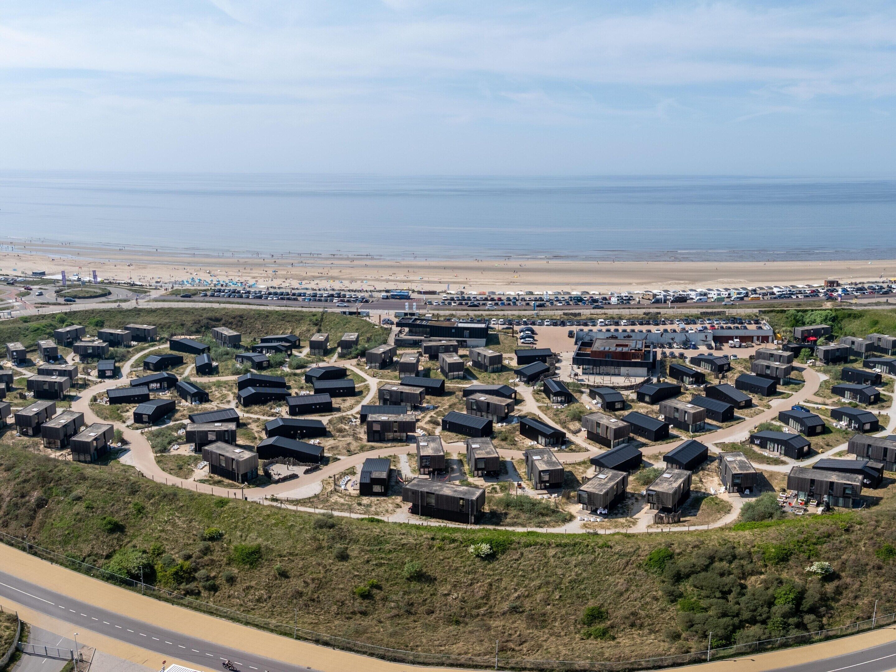 Vista aérea de una zona residencial costera con casas modernas de color oscuro dispuestas en círculo alrededor de una avenida central, con una playa y un estacionamiento al fondo.