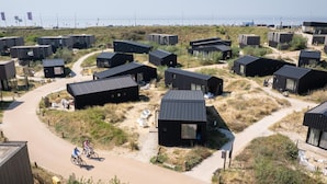 A coastal area with multiple modern, dark-colored cabins with flat roofs, arranged in a semi-circular pattern around a central path. - Special, Compound Group Accommodation, Near the Sea (Zandvoort)