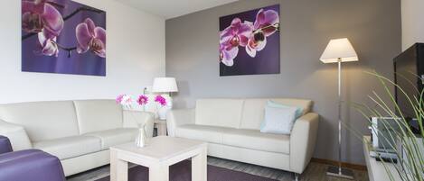 Uma sala de estar moderna com um sofá branco, poltronas roxas, uma mesa de centro e um abajur. Há duas pinturas de orquídeas nas paredes e um vaso com flores sobre a mesa de centro.