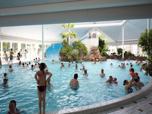 Ein Hallenbad mit mehreren Bahnen, einer Rutsche und einer Felsformation, umgeben von Palmen.
