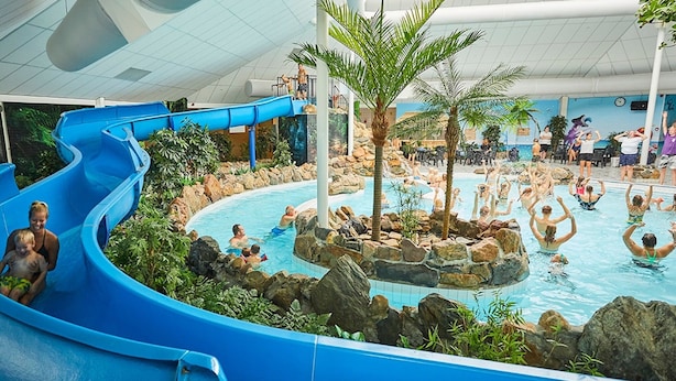 An indoor water park with a blue slide, multiple pools, and palm trees. - Villa With Garden in Holiday Park in Limburg (Roggel)