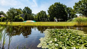A park with a pond, lily pads, a playground, and a house in the background. - Villa With Bubble Bath (Hellendoorn)