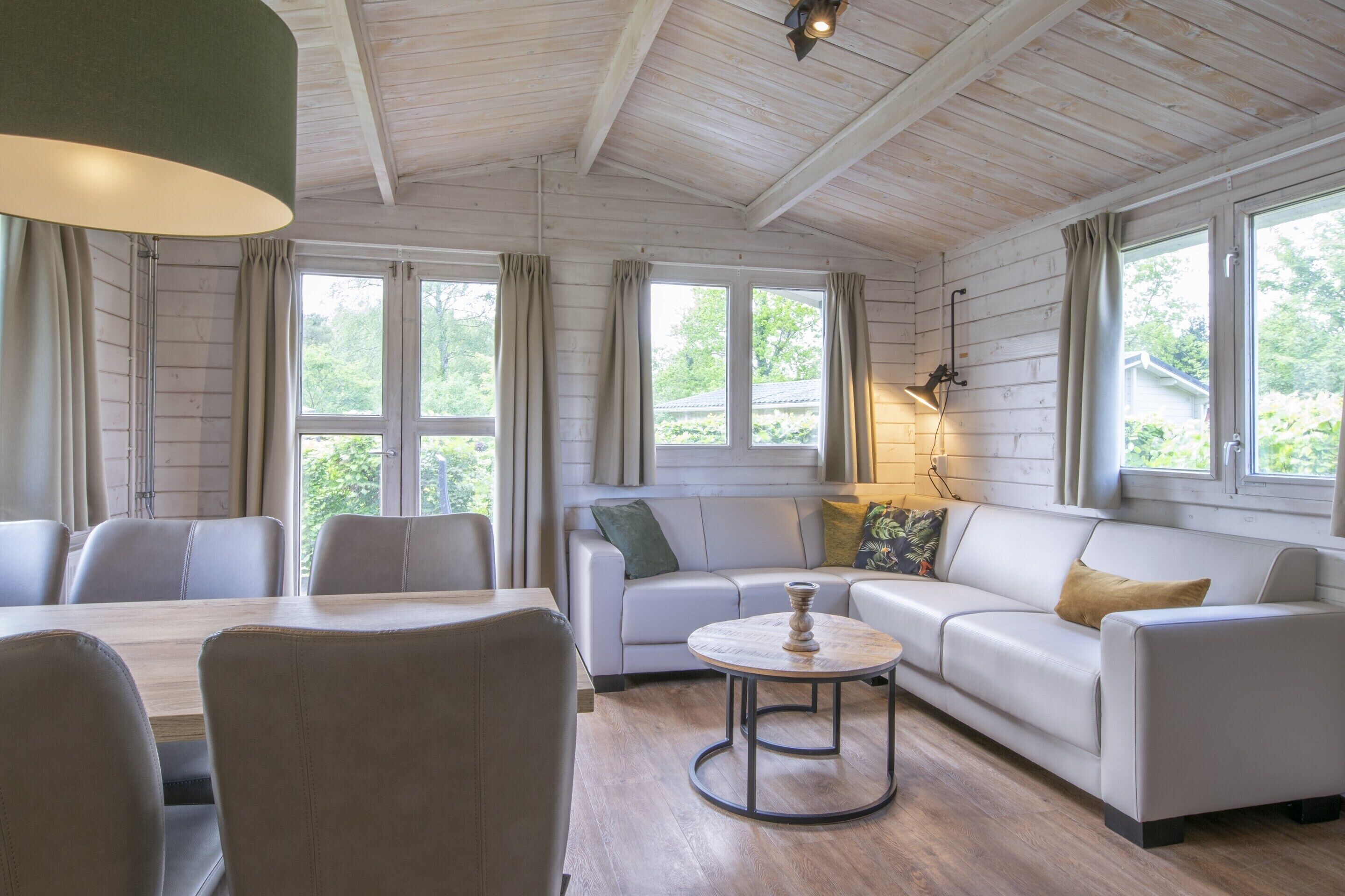 Une pièce comprenant une table à manger, des chaises, un canapé et une petite table basse ronde. On y trouve des fenêtres avec des rideaux, un ventilateur de plafond et une lampe fixée au mur.