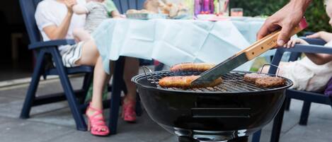 A barbecue grill with hamburgers being cooked, a table with a blue tablecloth, and people sitting at the table.