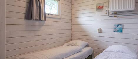 Quarto pequeno com uma cama de solteiro, paredes de madeira, janela com cortinas, um radiador e uma pequena imagem pendurada na parede.