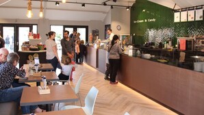 A modern café interior with wooden tables, white chairs, and a green wall with menu options. - Cozy Lodge With a Dishwasher in the Achterhoek (Winterswijk - Brinkheurne)