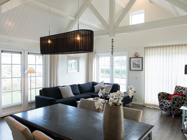 Relax in this airy living room with a sofa, armchair, and dining table, surrounded by large windows and a vase with fresh flowers.