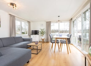 A modern living room with a sofa, a dining table with chairs, a TV stand, and large windows. - Modern and Cozy Chalet in Twente (Hoge Hexel)