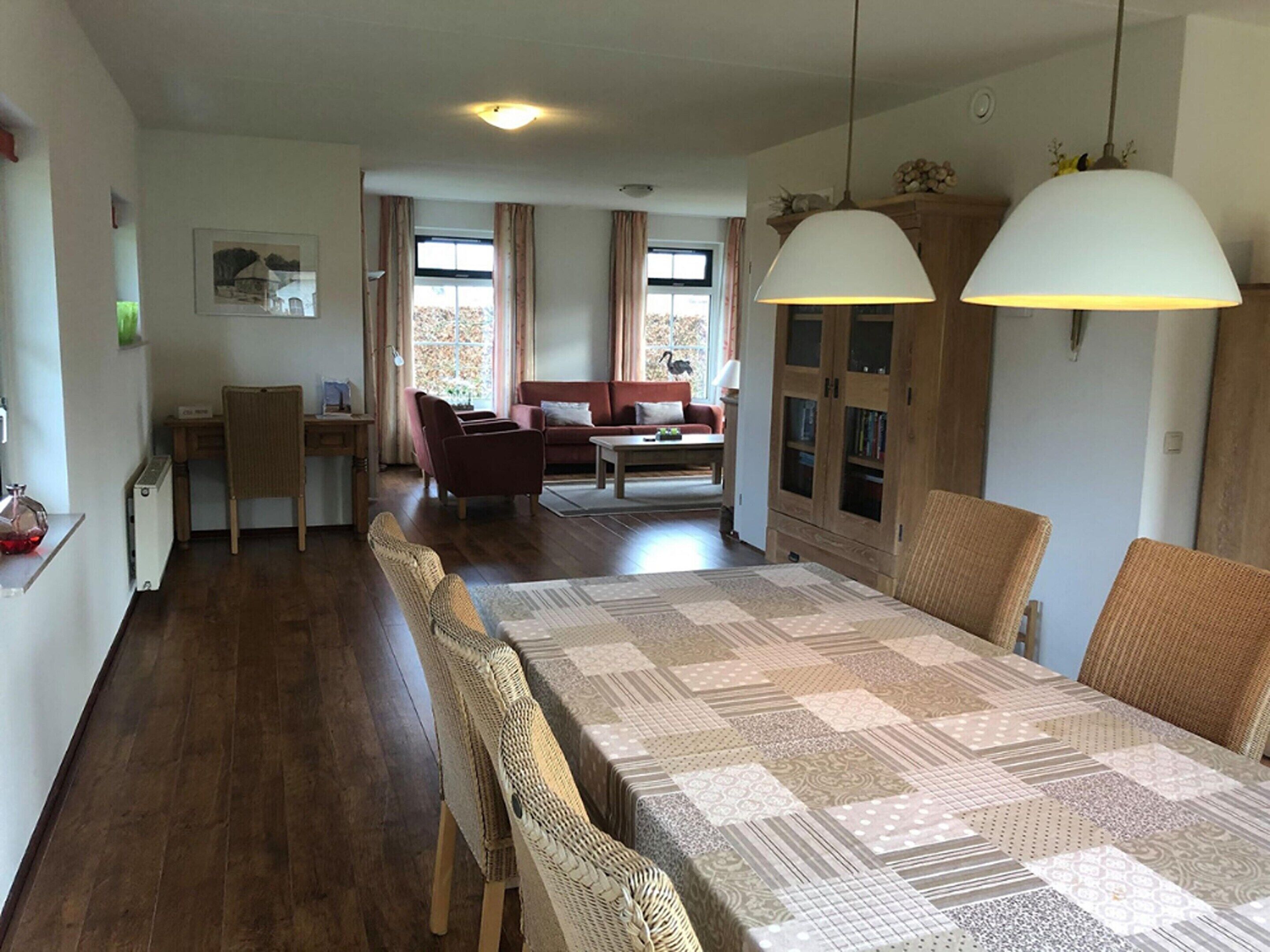 Een ruime woonruimte met een lange eettafel, een bank en een salontafel. Boven de tafel hangen twee hanglampen, en er is een boekenkast met boeken en decoratieve objecten.