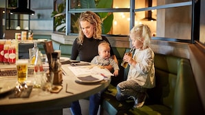 A woman and two children at a table in a restaurant with a modern interior, including a green couch and a window with plants. - Nice Villa With a Sauna in Limburg (Roggel)