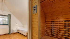 Ein gemütliches Zimmer mit schrägen Holzwänden, einem Bett, einem kleinen Nachttisch und einem Fenster mit Vorhängen.