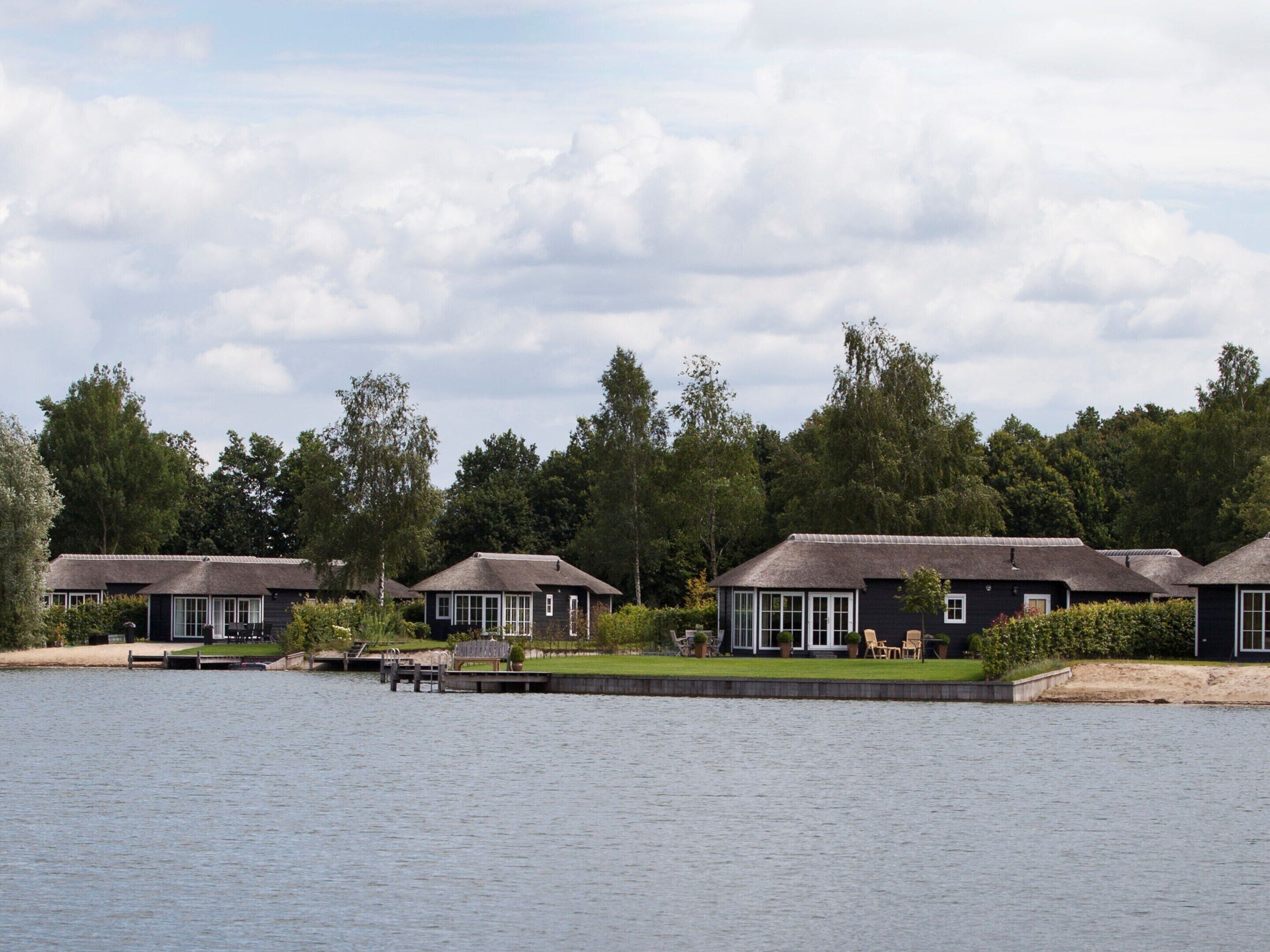 A row of dark-colored cottages with thatched roofs, situated by a calm lake, surrounded by lush greenery.