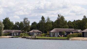 A row of dark-colored cottages with thatched roofs, situated by a calm lake, surrounded by lush greenery.