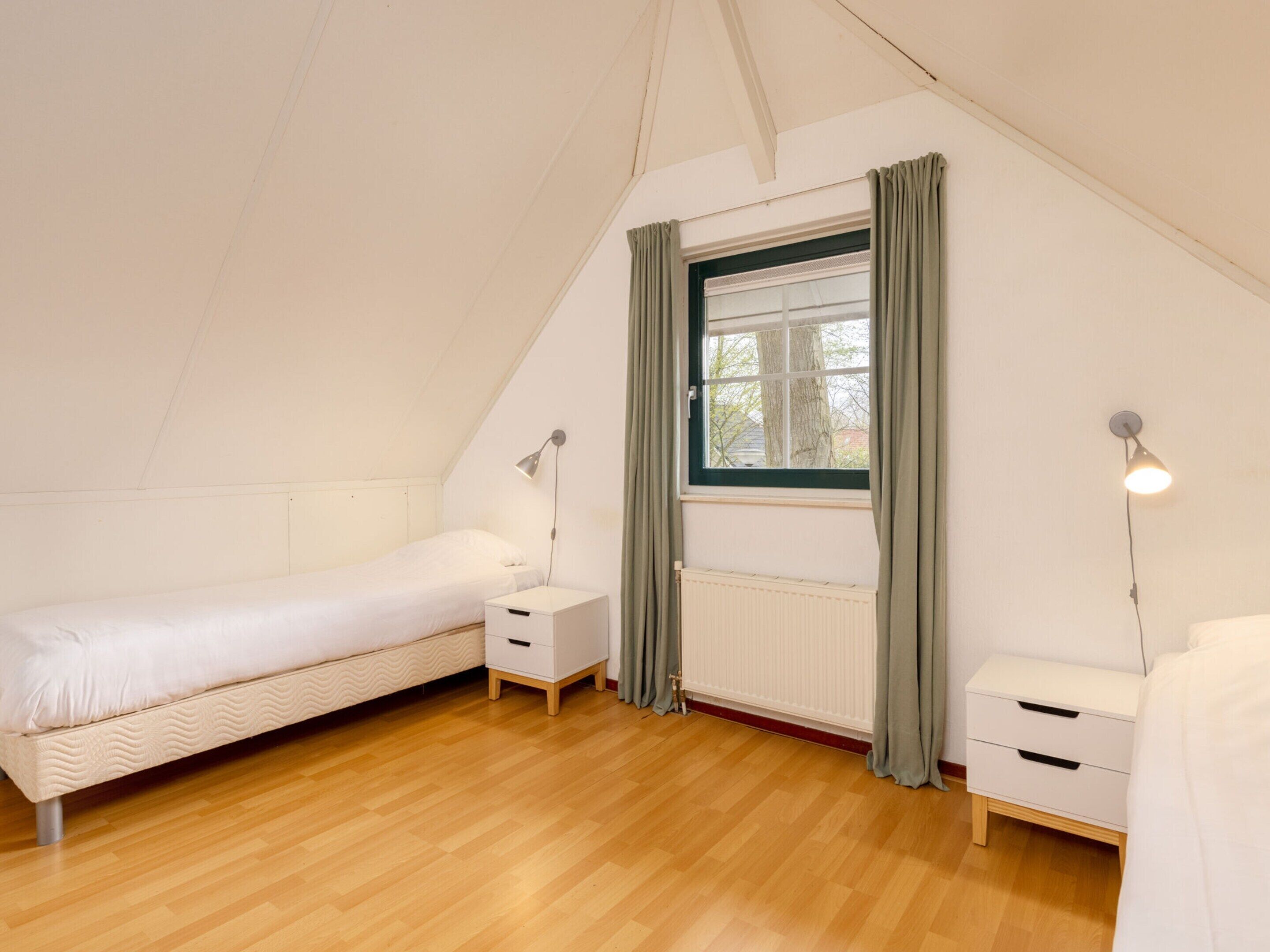A small, single-room with a sloped ceiling, a bed, two bedside tables, a window with curtains, and a radiator.