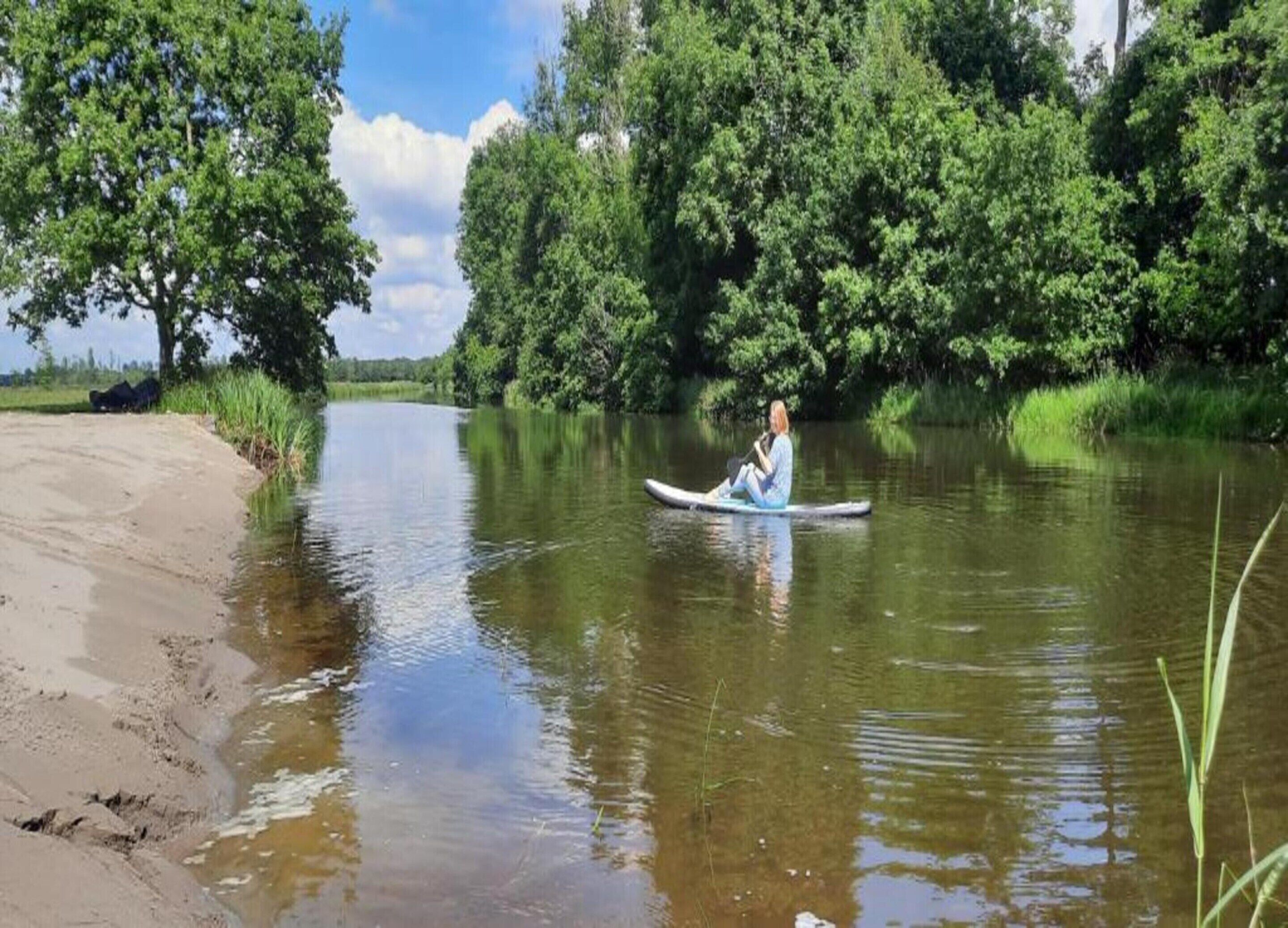 Une personne assise sur une planche à pagaie dans une rivière paisible, entourée d’une végétation luxuriante.