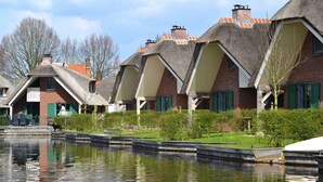A row of traditional thatched-roof cottages with brick walls and green shutters, situated beside a calm water body. - Stylish Thatched Villa with 2 Bathrooms near Giethoorn (Wanneperveen)