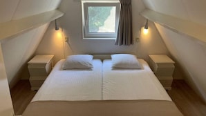 A twin bed in a room with a sloped ceiling, two bedside tables, a window with curtains, and two wall-mounted lamps. - Holiday House in Friesland With Garden (Sint Nicolaasga)
