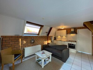 A compact living space with a loft bedroom, a kitchenette, a sofa, a small table, and a radiator.