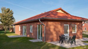A single-story brick building with a red-tiled roof, a patio with a table and chairs, and a clear sky.