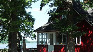A red wooden house with a white door and windows, surrounded by trees and a body of water.