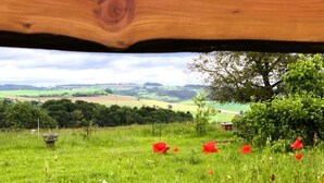 A view from under a wooden structure, overlooking a lush green field with scattered red flowers and a distant tree line. - Feriendorf Reinskopf in der Eifel (Schoenecken)