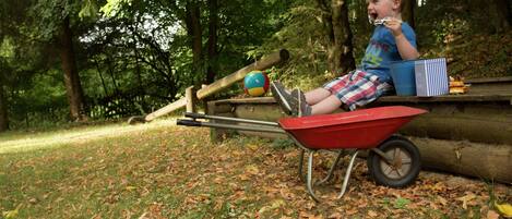 A child sitting in a red wheelbarrow in a park with trees and a playground in the background.