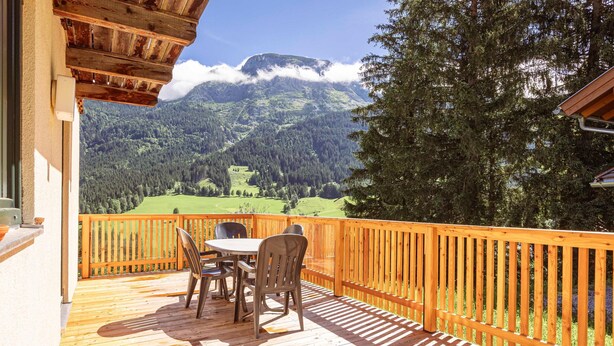 A wooden deck with a table and chairs, overlooking a mountainous landscape with a wooden railing.