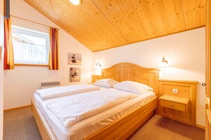 Un dormitorio con techo de madera, una cama con ropa de cama blanca, una mesita de noche, una lámpara de pared y una ventana con cortinas.