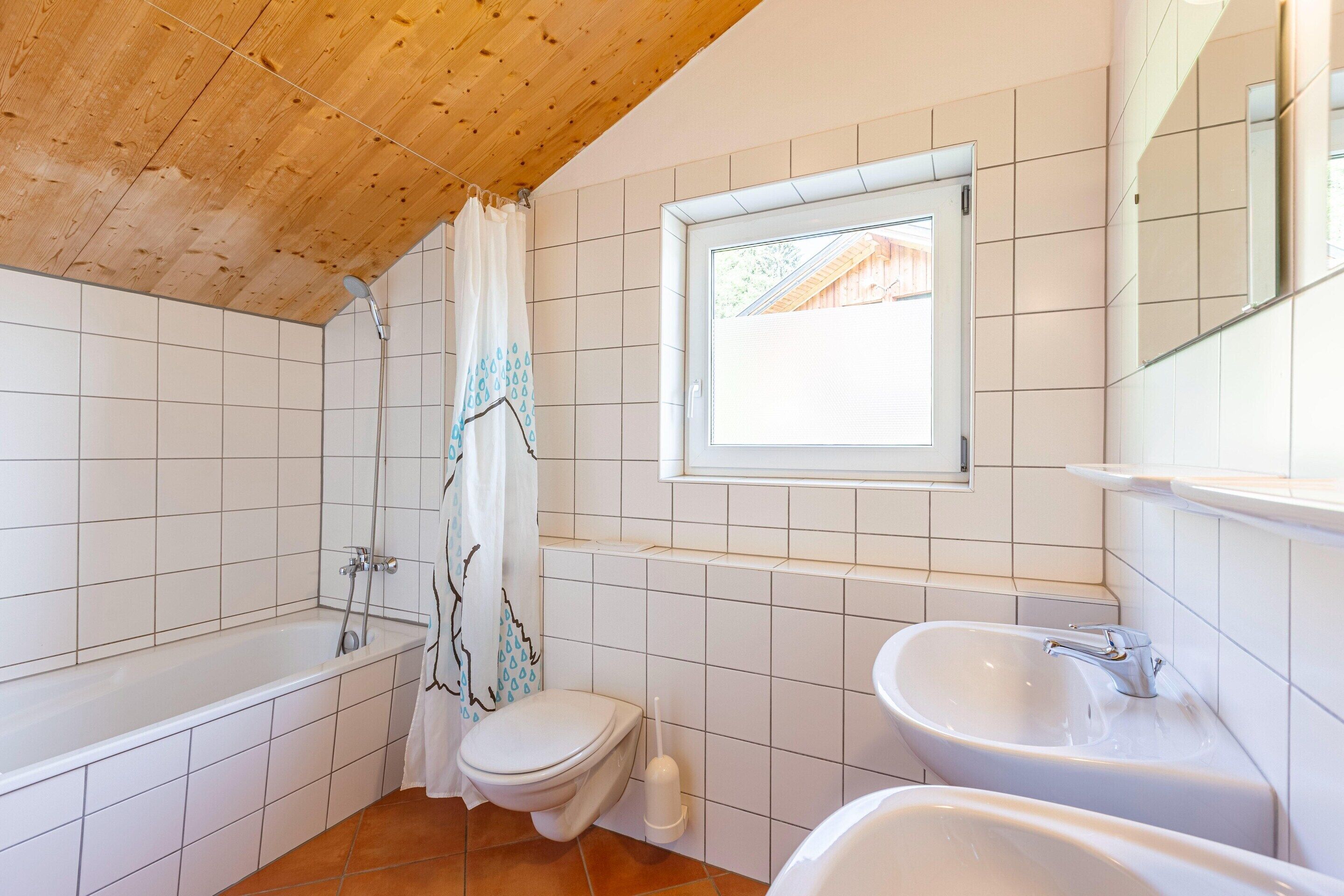 A bathroom with a wooden ceiling, white tiled walls, a window, a bathtub, a toilet, and a sink.