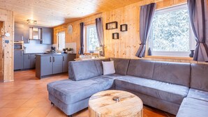 A modern log cabin interior with a grey sectional sofa, wooden flooring, and a kitchen area.