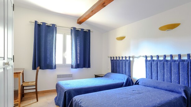 A room with two beds, a chair, a table, a window with curtains, and a radiator. - Holiday Home in Bormes-les-mimosas Near Sea (Bormes-Les-Mimosas)