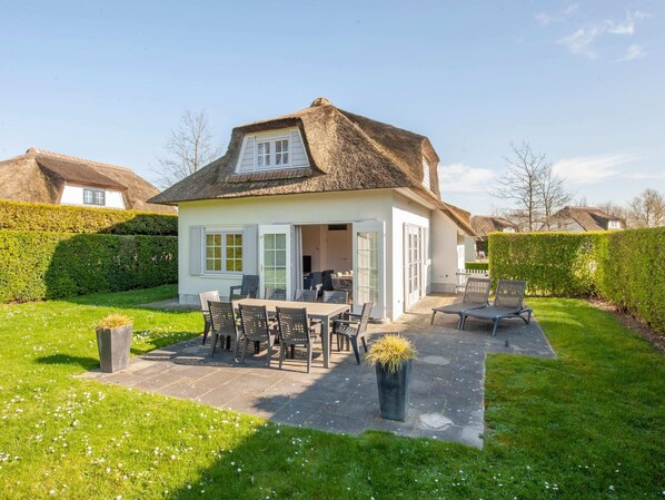 Une maison avec un toit de chaume, une terrasse avec une table et des chaises, et un jardin bien entretenu.